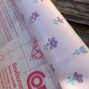 May include: A roll of floral patterned contact paper. The paper has a light pink background with repeating designs of purple, blue, and pink flowers. The paper is partially unrolled, revealing a red grid pattern and text in French.