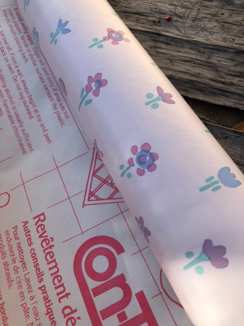 May include: A roll of floral patterned contact paper. The paper has a light pink background with repeating designs of purple, blue, and pink flowers. The paper is partially unrolled, revealing a red grid pattern and text in French.