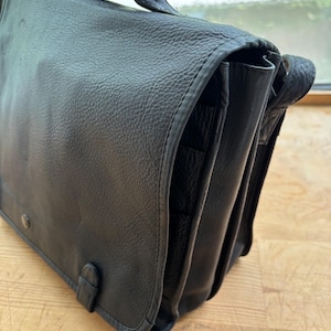 Puede incluir: Bolso mensajero de cuero negro con asa y cierre de solapa. El bolso tiene una superficie texturizada y costuras visibles en los bordes. Cuenta con una hebilla y un cierre a presi&oacute;n. El bolso est&aacute; sobre una superficie de madera.