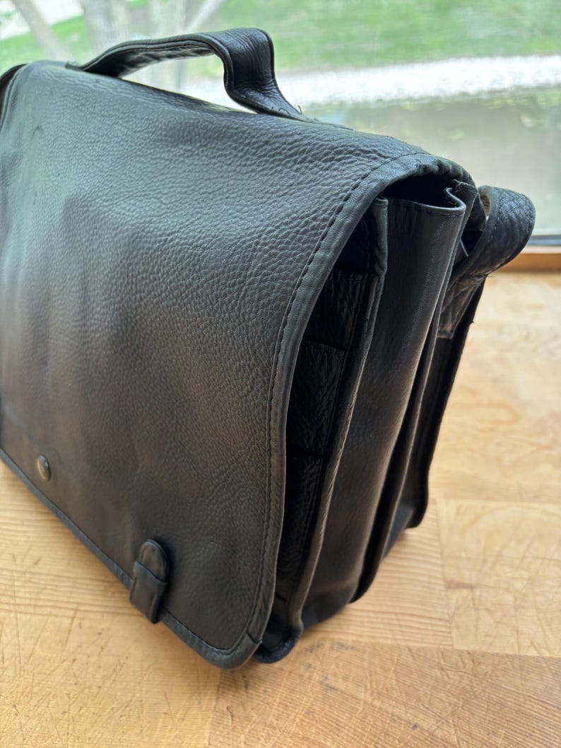 Puede incluir: Bolso mensajero de cuero negro con asa y cierre de solapa. El bolso tiene una superficie texturizada y costuras visibles en los bordes. Cuenta con una hebilla y un cierre a presi&oacute;n. El bolso est&aacute; sobre una superficie de madera.