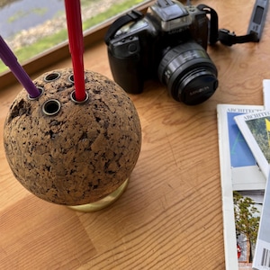 May include: A spherical cork pen holder with three holes, holding a red and purple pen. A black Minolta camera with a lens cap and strap sits behind the pen holder. Architectural magazines are visible on a wooden surface.