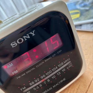 May include: A vintage SONY Dream Machine digital clock radio. The clock displays 3:15 in red numerals. The radio has AM/FM capabilities and a black face with white lettering. The model number is ICF-C121.