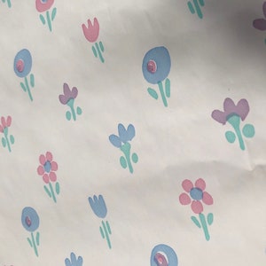 May include: White background with a repeating pattern of stylized flowers in pink, blue, and purple. The flowers have green stems and leaves. The design is simple and whimsical.