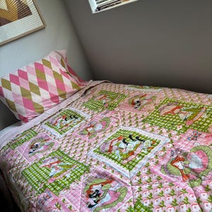 May include: A quilted bedspread featuring a patchwork design of pink and green checkered squares, floral patterns, and illustrations of figures with red hair. A matching pillowcase with a pink and olive green diamond pattern sits at the head.