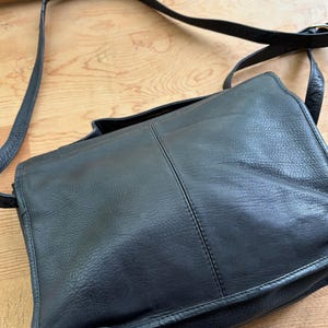 Puede incluir: Bolso mensajero de cuero negro con una correa larga ajustable. El bolso tiene un cierre de solapa y una forma rectangular. El cuero parece ligeramente texturizado. El bolso est&aacute; dise&ntilde;ado para llevarse al hombro.