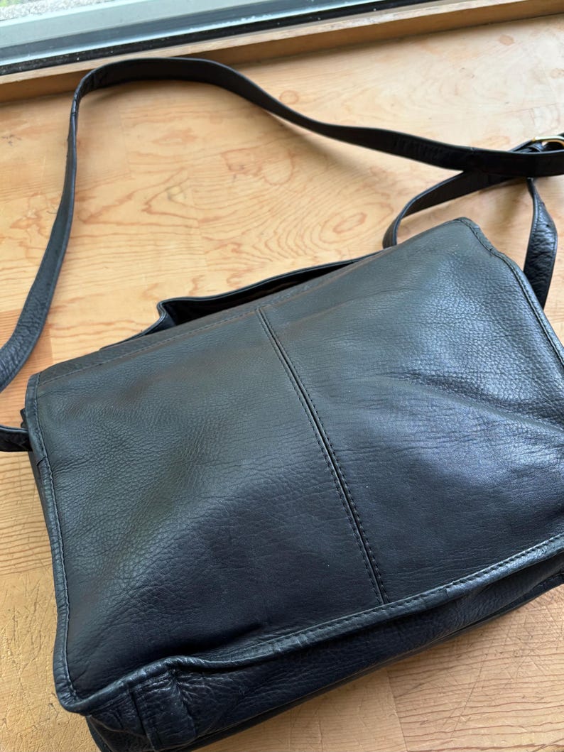 Puede incluir: Bolso mensajero de cuero negro con una correa larga ajustable. El bolso tiene un cierre de solapa y una forma rectangular. El cuero parece ligeramente texturizado. El bolso est&aacute; dise&ntilde;ado para llevarse al hombro.