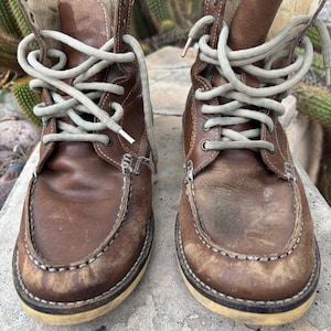 May include: A pair of brown leather boots with light-colored laces. The boots have a moccasin-style toe and a cream-colored sole. The leather shows signs of wear, with scuff marks and a distressed appearance. The boots are laced up and ready to wear.