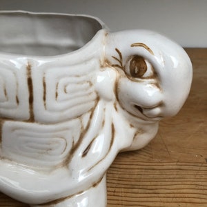 Puede incluir: Maceta de cer&aacute;mica blanca en forma de tortuga con detalles pintados en marr&oacute;n. La tortuga tiene una cara sonriente y se ve un ojo. La maceta est&aacute; vac&iacute;a.