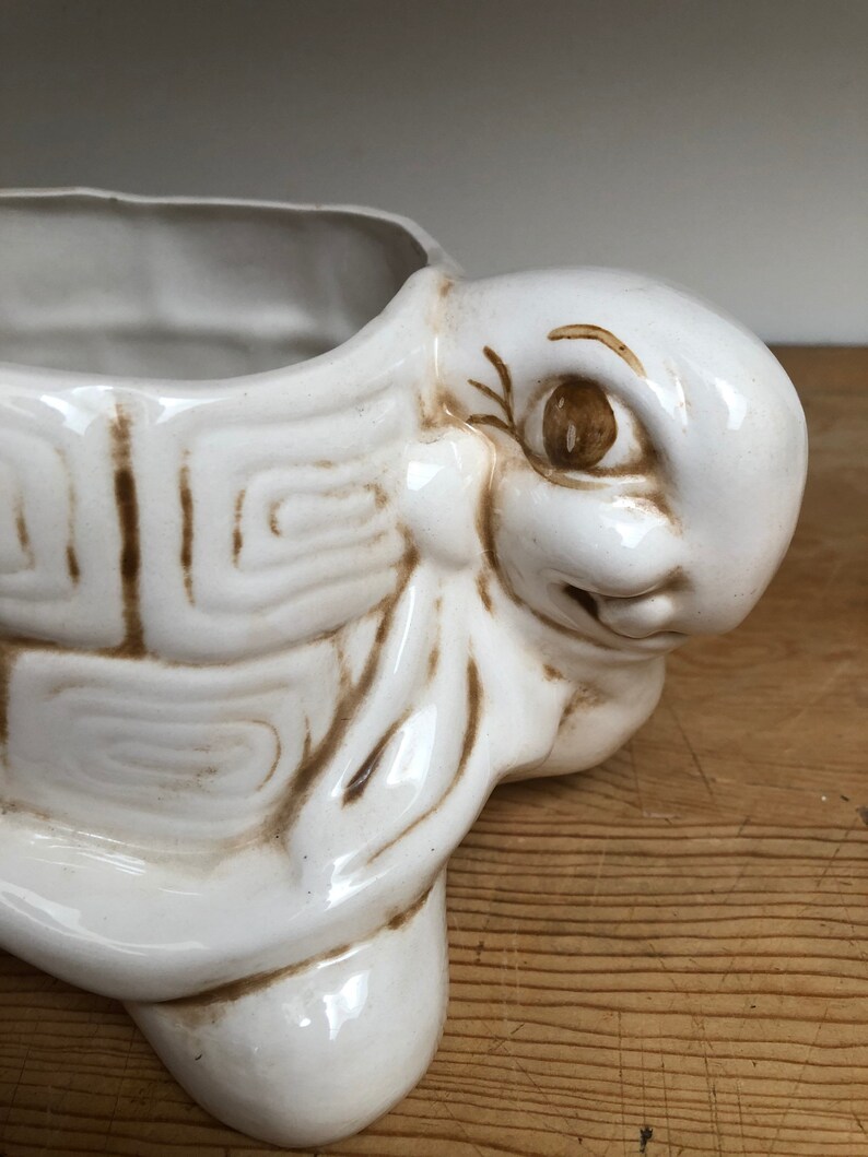 Puede incluir: Maceta de cer&aacute;mica blanca en forma de tortuga con detalles pintados en marr&oacute;n. La tortuga tiene una cara sonriente y se ve un ojo. La maceta est&aacute; vac&iacute;a.