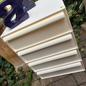 May include: A white plastic storage unit featuring four stacked drawers with rounded horizontal handles. A dark purple ceramic letter A is positioned on the top surface of the unit. The storage organizer has a clean, modular design with a smooth finish.
