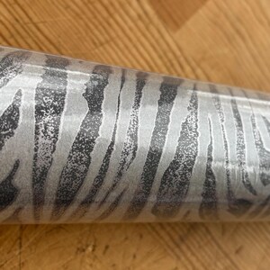 May include: A roll of wallpaper featuring a zebra stripe pattern in shades of gray and silver. The design has a textured appearance, with dark gray stripes against a lighter silver background. The roll is cylindrical.