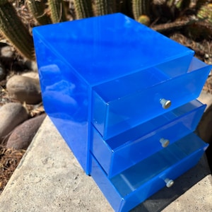 May include: A bright blue translucent acrylic storage unit featuring three stacked drawers. Each drawer is equipped with a small, round, light-colored knob for opening. The unit has a smooth, glossy finish and a rectangular shape with clean edges.