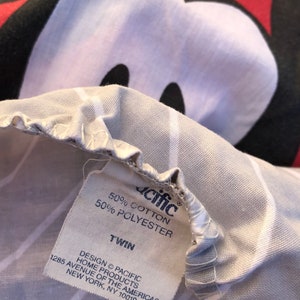 May include: A white twin-sized bed sheet with a Mickey Mouse print. The sheet has a white and gray striped pattern. The sheet is made of 50% cotton and 50% polyester. The label reads "Pacific Home Products, 1285 Avenue of the Americas, New York, NY 10019."