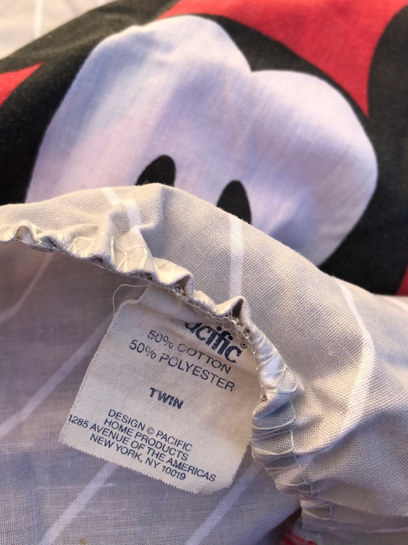 May include: A white twin-sized bed sheet with a Mickey Mouse print. The sheet has a white and gray striped pattern. The sheet is made of 50% cotton and 50% polyester. The label reads "Pacific Home Products, 1285 Avenue of the Americas, New York, NY 10019."