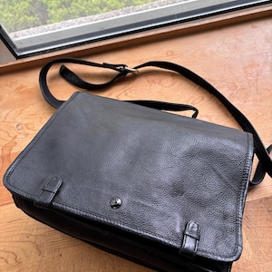 May include: Black leather messenger bag with a flap closure, adjustable shoulder strap, and two buckle closures on the front. The bag is sitting on a wooden surface.