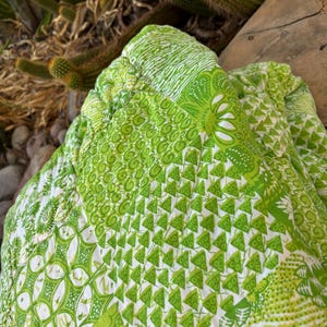 May include: A folded green and white patterned fabric with various designs, including floral and geometric shapes. The fabric is a vibrant shade of green, with white accents. The fabric is likely a quilt or a similar textile.
