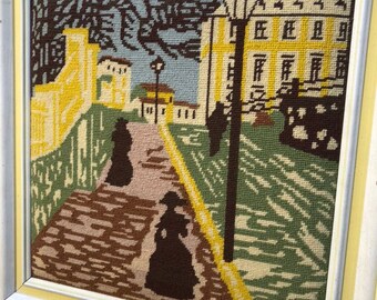 May include: A framed needlepoint artwork depicting a street scene with two figures in black dresses walking down a cobblestone path. The scene is set in front of a yellow building with a green lawn and trees.