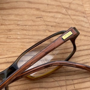 May include: Close-up of eyeglasses with brown frames and a gold accent on the temple. The lenses are clear, and the arms of the glasses are a gradient of brown and yellow.