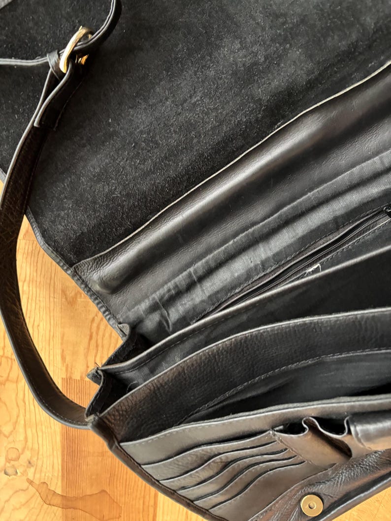 Puede incluir: Bolso de hombro de cuero negro con m&uacute;ltiples compartimentos y una correa larga. El bolso presenta una hebilla dorada y un cierre a presi&oacute;n. El interior est&aacute; forrado con un material suave y oscuro.