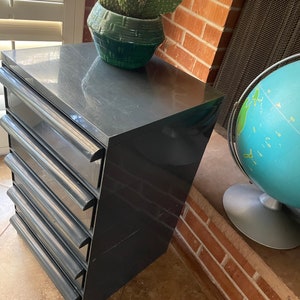 May include: A dark gray plastic drawer unit with five drawers. The drawer unit is sitting on a tiled floor. A potted cactus plant is sitting on top of the drawer unit.