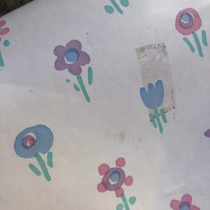 May include: White paper with a repeating pattern of stylized flowers in shades of purple, blue, and pink. The flowers have green stems and leaves. The paper has some discoloration and a piece of tape.