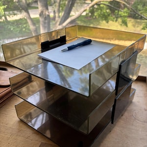May include: A three-tier desk organizer made of translucent amber-colored plastic with black side supports. The top tray holds a single sheet of white paper and a dark blue ballpoint pen. The structure features an open-front design for documents.
