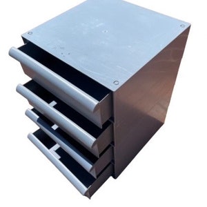 May include: A gray metal drawer unit with four drawers. The drawers are all closed and the unit is sitting on a white surface.
