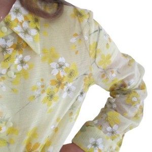 May include: A sheer yellow blouse with a floral print featuring white and yellow flowers. The blouse has a collared neckline and long sleeves.