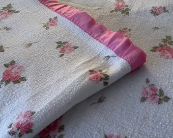 May include: A white blanket with a pink floral pattern. The blanket has a pink satin trim around the edges.