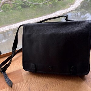 Puede incluir: Bolso mensajero de cuero negro con cierre de solapa, asa superior y bandolera ajustable. El bolso tiene dos cierres de hebilla en la parte delantera y un peque&ntilde;o logotipo. El bolso est&aacute; sobre una superficie de madera.