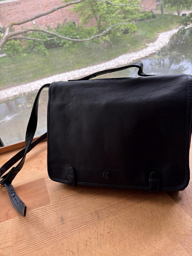 Puede incluir: Bolso mensajero de cuero negro con cierre de solapa, asa superior y bandolera ajustable. El bolso tiene dos cierres de hebilla en la parte delantera y un peque&ntilde;o logotipo. El bolso est&aacute; sobre una superficie de madera.