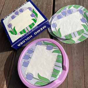 May include: A set of purple memo holders with a floral design featuring white tulips with green leaves. The holders are made of plastic and have a white memo pad inside.