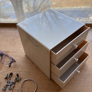 May include: A cream-colored jewelry box with three pull-out drawers. The box is rectangular with a slightly glossy finish. A pair of glasses, earrings, a bracelet, and a ring are next to the box on a wooden surface.