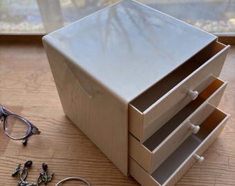 Puede incluir: Una joyero de color crema con tres cajones extraíbles. La caja es rectangular con un acabado ligeramente brillante. Un par de gafas, pendientes, una pulsera y un anillo están al lado de la caja sobre una superficie de madera.