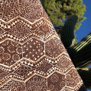 May include: Close-up of a folded fabric with a repeating geometric pattern. The fabric features a dark brown background with lighter brown and beige hexagonal shapes filled with various designs, including circles, dots, and floral motifs. The fabric is set against a backdrop of a blue sky, a green tree, and a cactus.