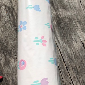 May include: Rolled white paper with a repeating floral pattern in pastel colors. The design features stylized flowers in shades of pink, blue, and purple, with green accents. The paper is likely for wrapping or crafting.