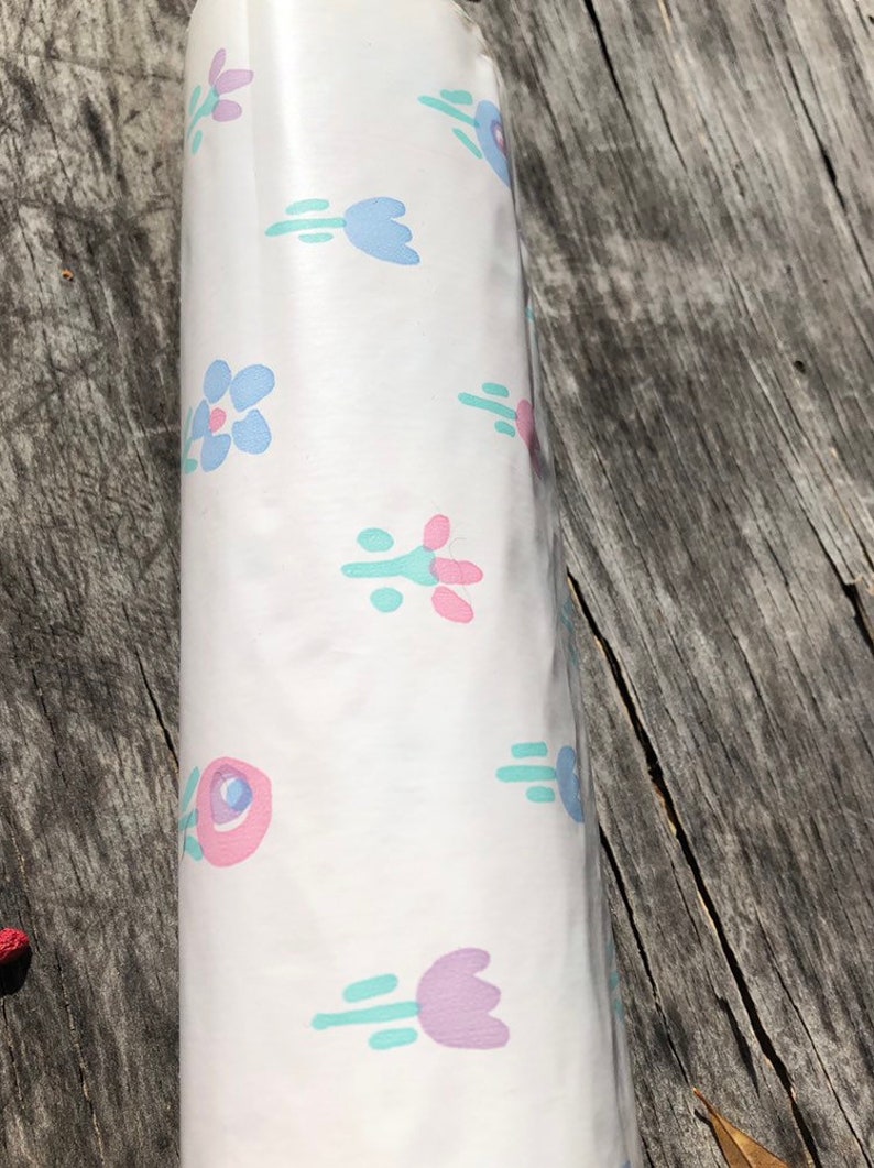 May include: Rolled white paper with a repeating floral pattern in pastel colors. The design features stylized flowers in shades of pink, blue, and purple, with green accents. The paper is likely for wrapping or crafting.