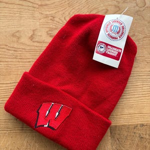 May include: A red knit beanie with a folded brim. The beanie features a sewn-on patch with a white and black "W" logo. A white tag with collegiate licensing information is attached.