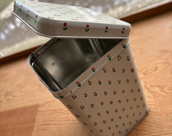 Puede incluir: Una lata de almacenamiento rectangular de metal con tapa abatible, que presenta un color base blanco roto decorado con un patrón repetitivo de pequeños tulipanes rojos con tallos y hojas verdes. El interior del recipiente es plateado.