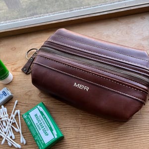 May include: A brown leather toiletry bag with the initials "MBR" embossed on the front. The bag has a zipper closure and a small leather pull tab. Beside the bag are a green bar of soap, cotton swabs, and a tube of toothpaste.