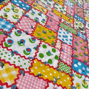 May include: A colorful patchwork quilt with a variety of patterns, including flowers, polka dots, and gingham. The quilt is made with a white background and features blue, pink, yellow, and green colors. The edges of the quilt are finished with a blue fringe.