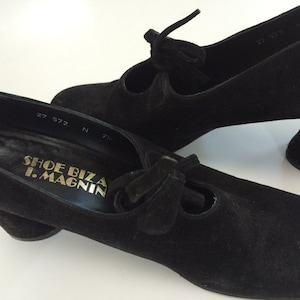 May include: A pair of black suede shoes with a chunky heel and a bow detail. The shoes are labelled "Shoe Biz by I. Magnin" and have a size tag that reads "27 572 N 7 1/2".
