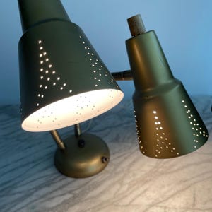 May include: Two olive green vintage desk lamps with perforated shades. The lamps have a brass base and wooden accents. The shades are cone-shaped and have small holes that emit light. The lamps are angled towards the viewer.