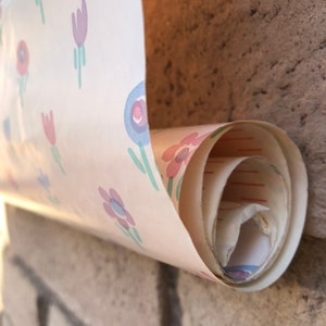 May include: A roll of vintage wallpaper with a repeating floral pattern in pink, purple, and blue on a cream background. The wallpaper is partially unrolled, revealing the rolled-up interior. The wallpaper is against a brick wall.