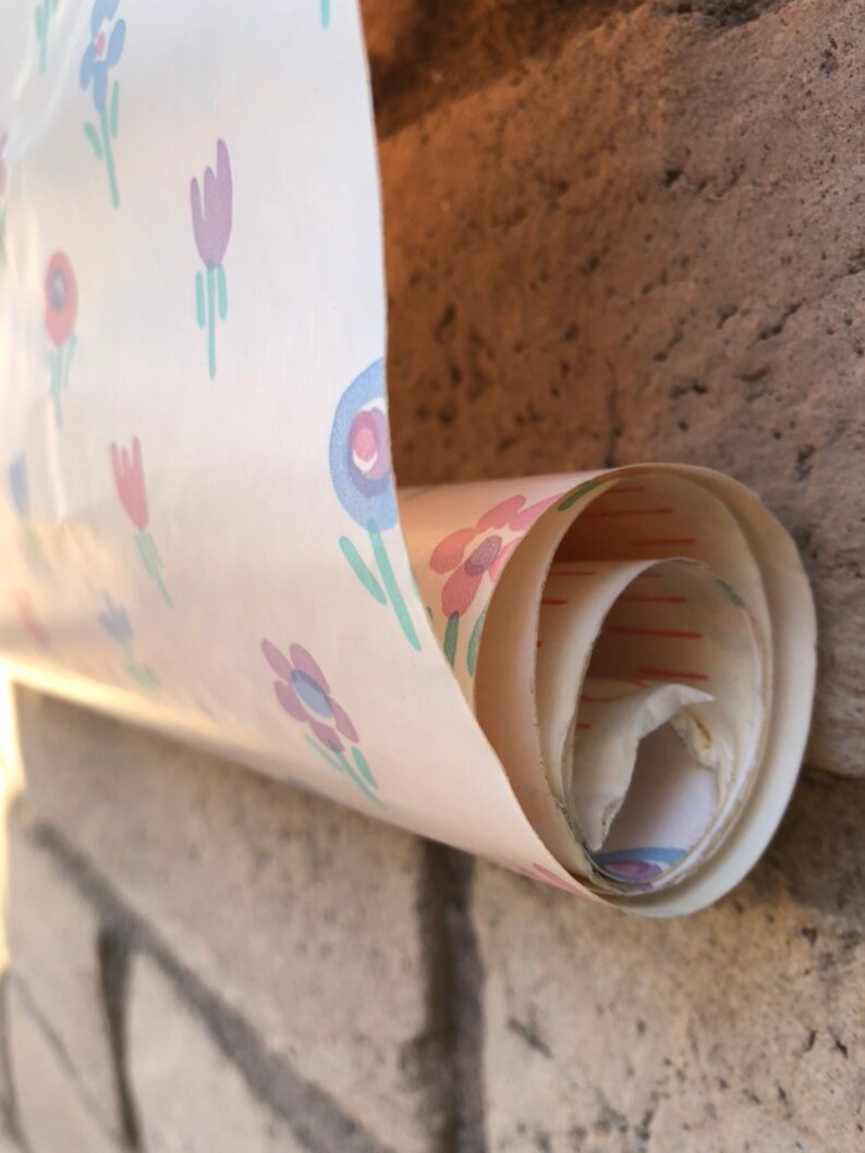 May include: A roll of vintage wallpaper with a repeating floral pattern in pink, purple, and blue on a cream background. The wallpaper is partially unrolled, revealing the rolled-up interior. The wallpaper is against a brick wall.