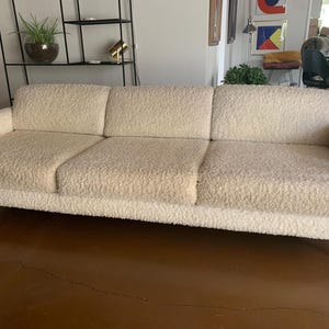May include: A three-cushion sofa upholstered in a cream-colored, textured bouclé fabric. The sofa features a long, rectangular design with straight back and seat cushions, supported by a metallic, silver-toned frame visible at the armrests.