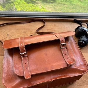 May include: A brown leather messenger bag with a flap closure and adjustable shoulder strap. The bag features two buckle straps and white stitching. A camera is visible in the background.
