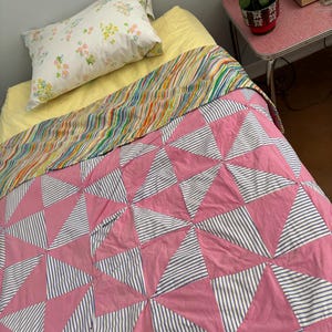 May include: A bed with a pink and white quilt, a yellow sheet, a pillow with a floral pattern, and a striped blanket. The quilt features a pinwheel design with pink triangles and blue and white striped triangles. The bed is in a room with a small side table.
