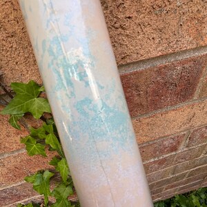 May include: A rolled-up wallpaper roll with a light beige base and irregular turquoise and white patterns. The roll is positioned diagonally against a red brick wall, with green ivy leaves in the foreground.