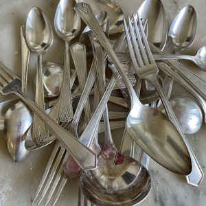 May include: A pile of silver silverware, including spoons, forks, and a serving spoon, all arranged on a marble surface.